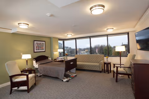 A spacious senior living room with a hospital-style bed, two armchairs, a sofa, side tables with lamps, a large window showing an outdoor view, and a wall-mounted television.