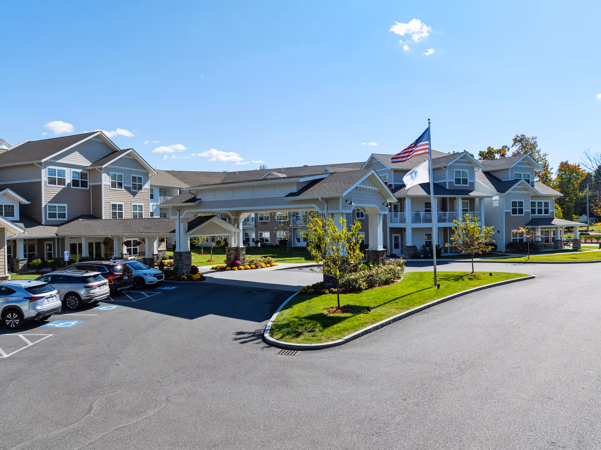 Exterior view of Kennedy Meadows Gracious Retirement Living facility on a sunny day, showing a large building with multiple windows, a covered entrance with stone pillars, a flagpole with American and state flags, landscaped greenery, and a parking lot with several cars.