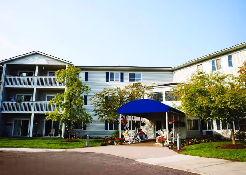 Exterior view of a multi-story senior living facility with white siding and blue shutters. The entrance features a blue canopy with several people sitting on white rocking chairs on the porch. There are trees and well-maintained landscaping around the building under a clear blue sky.
