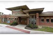 Exterior view of a single-story brick building with large windows and a covered entrance, identified as the Daughters of Miriam Center for Nursing and Rehabilitation. The building is surrounded by a sidewalk and some landscaping with bushes and grass.