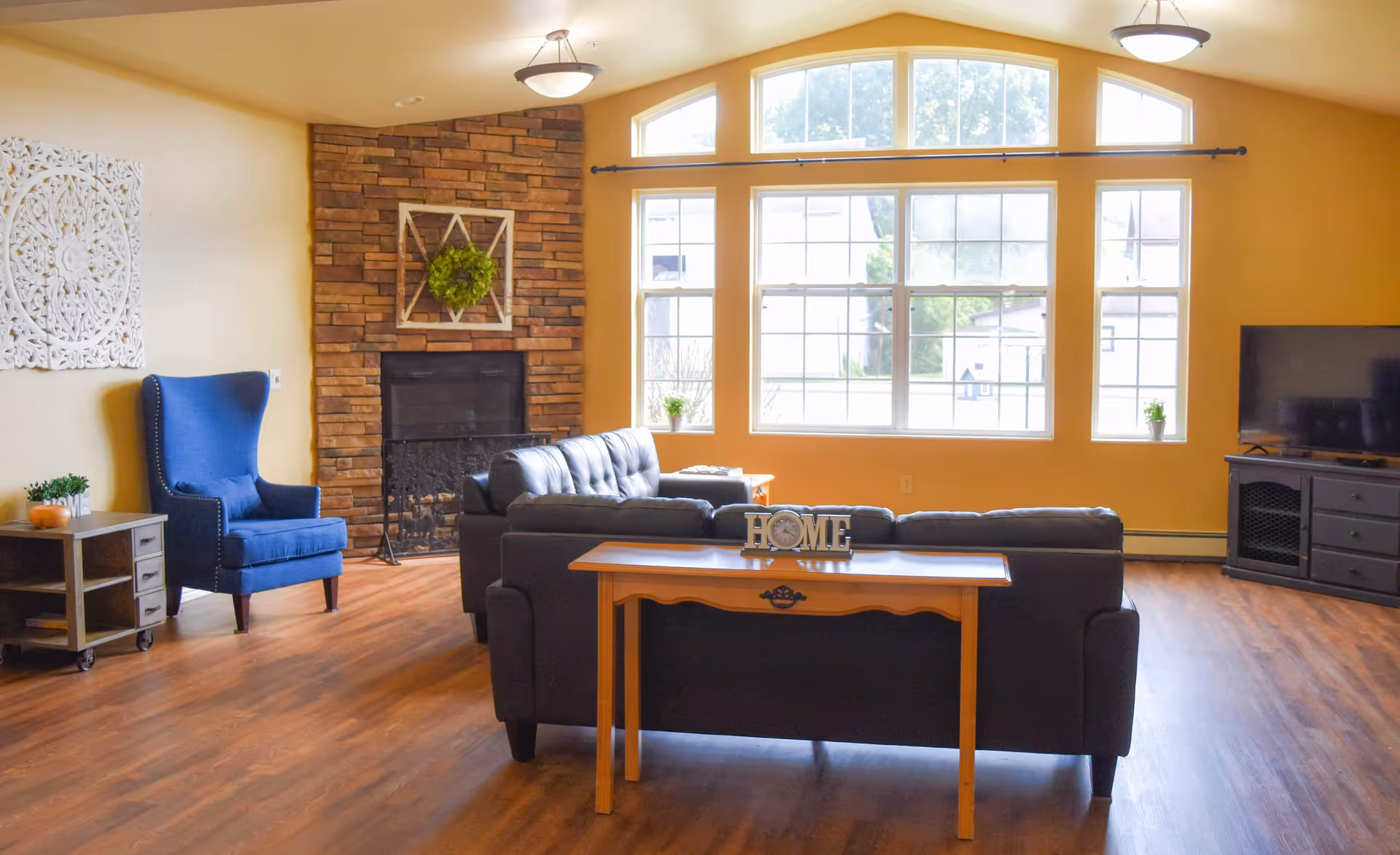 A cozy living room with a large window letting in natural light. The room features a dark leather sofa, a wooden table with a decorative 'HOME' sign, a blue armchair, a side table with a small plant and pumpkin decoration, a brick fireplace with a green wreath, and a TV on a black stand. The walls are painted yellow and the floor is wooden.