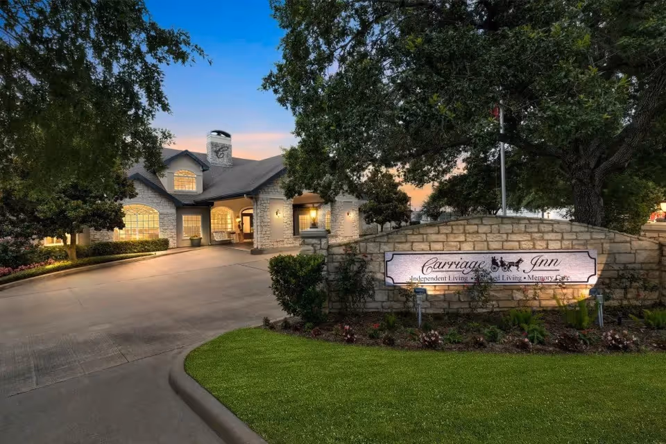 Exterior view of Carriage Inn Katy senior living facility at dusk, showing a stone building with warm lights inside, surrounded by trees and landscaping, with a stone sign in front displaying the facility's name and services.