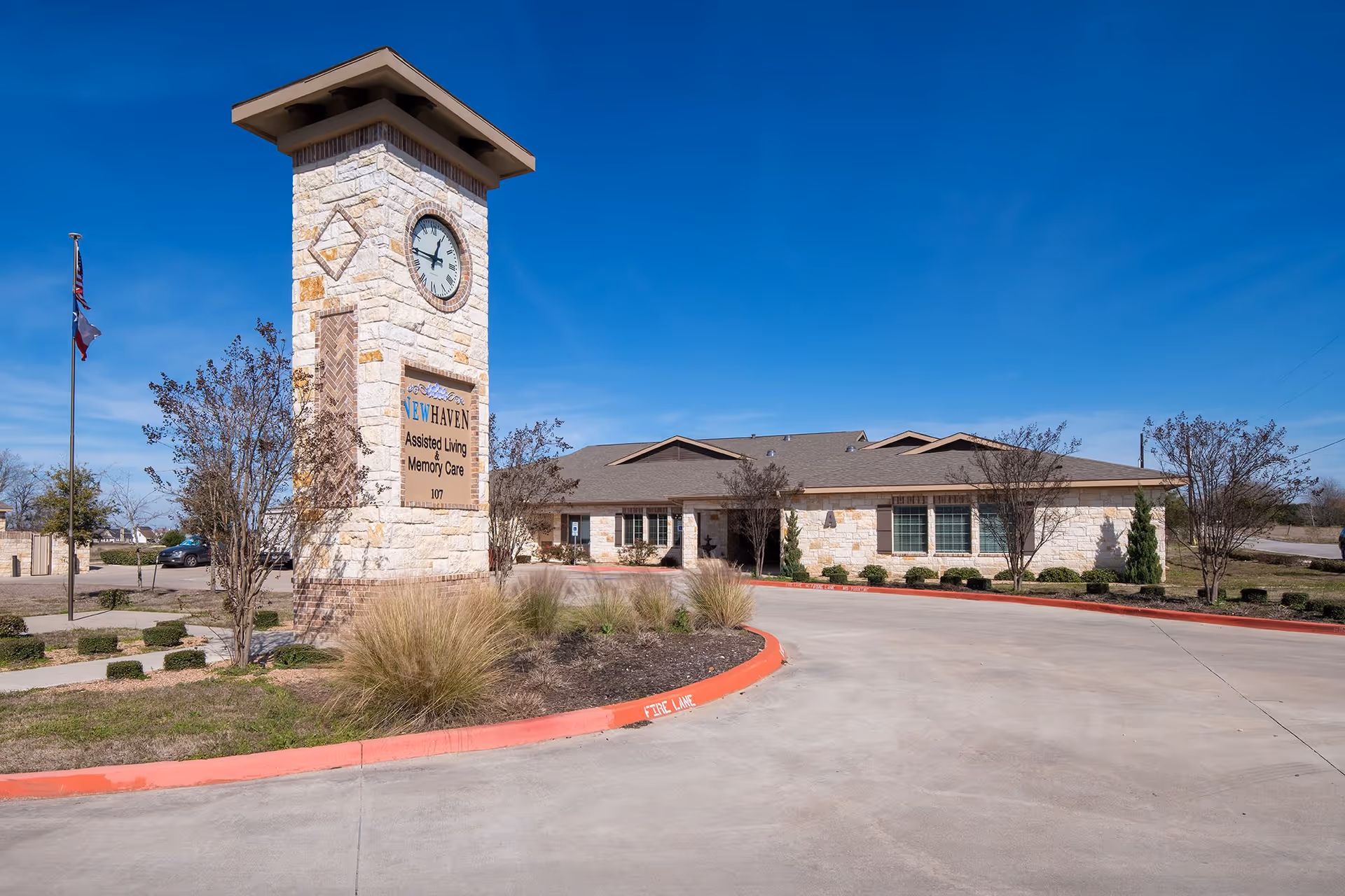 Exterior view of New Haven Assisted Living and Memory Care of Kyle facility on a clear day, featuring a stone clock tower with the facility's name and a single-story building surrounded by landscaping and a driveway.
