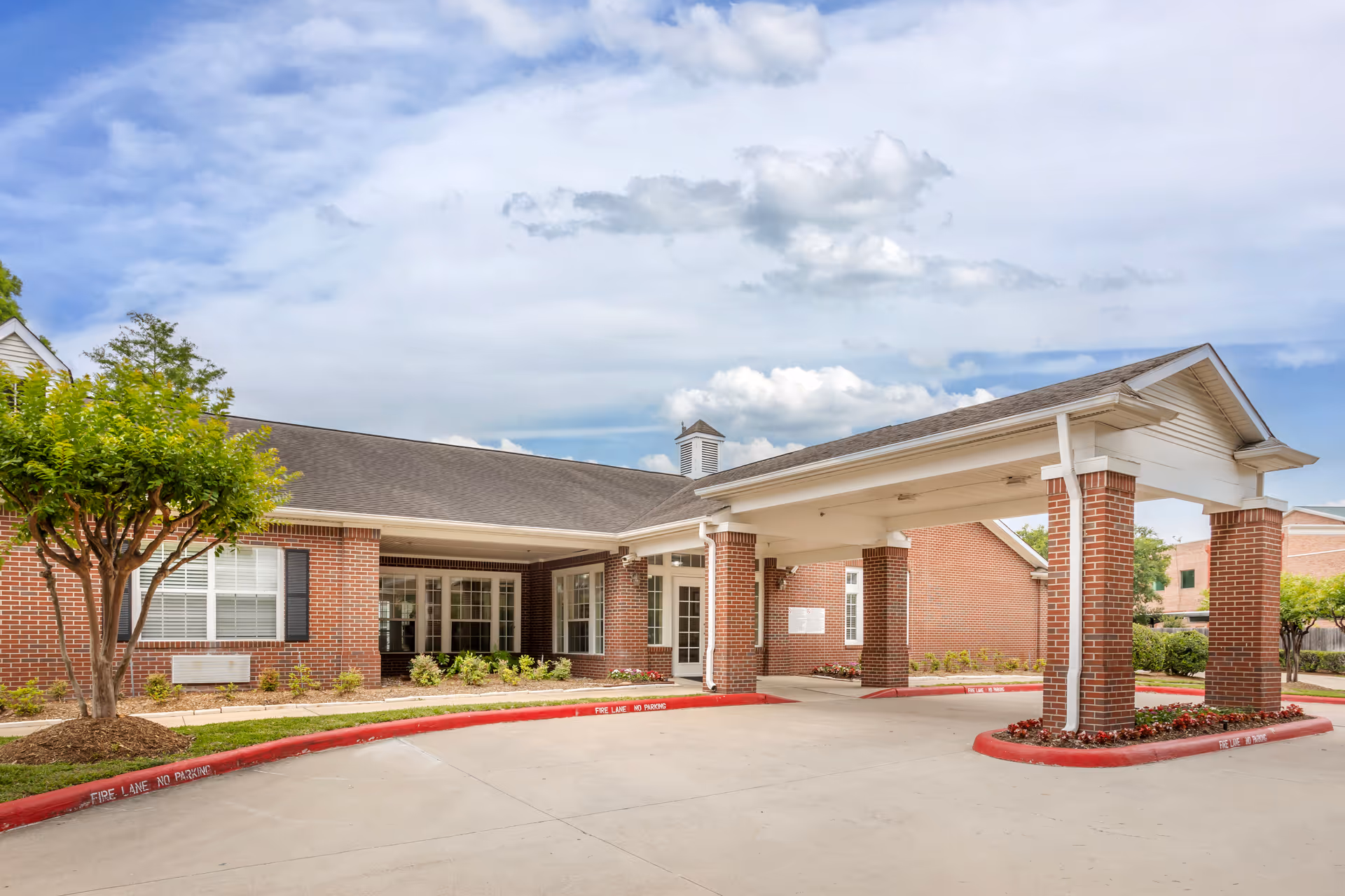 Exterior view of a single-story brick building with a covered entrance supported by brick columns. The building has white trim and a dark shingled roof. There are small landscaped areas with bushes and flowers around the building, and a tree on the left side. The sky is partly cloudy.