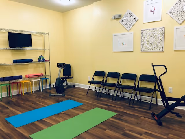 A small exercise room with two yoga mats on a wooden floor, a stationary exercise bike, a row of five black folding chairs against a yellow wall, colorful stools under a metal shelving unit holding small weights and rolled-up mats, and framed artwork on the walls.
