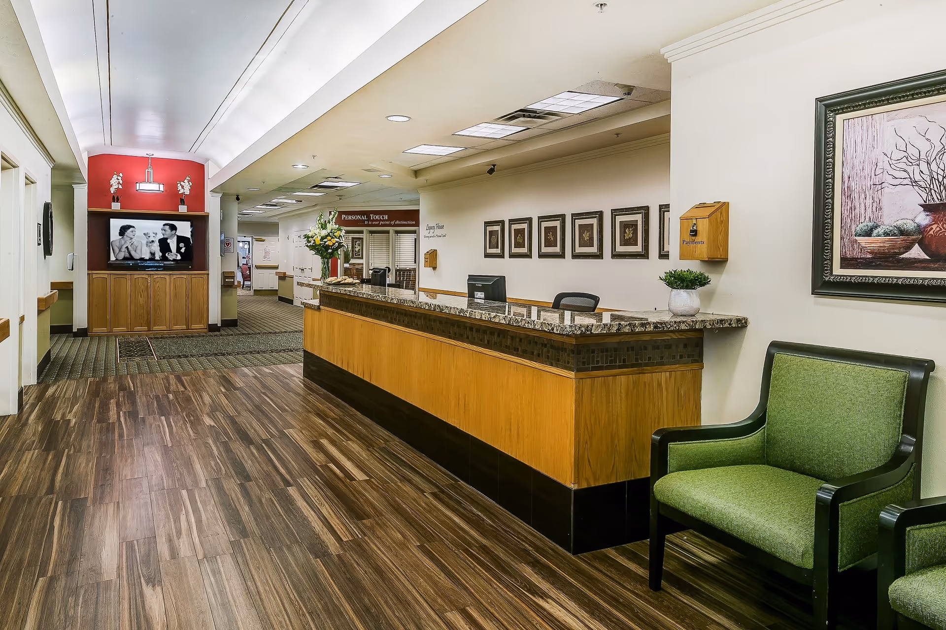 Reception area of Legacy House of Taylorsville featuring a wooden front desk with granite countertop, a green upholstered chair, framed artwork on the walls, and a hallway leading to other rooms. There is a TV mounted on a wooden cabinet in the background showing a black and white image of a man and woman.