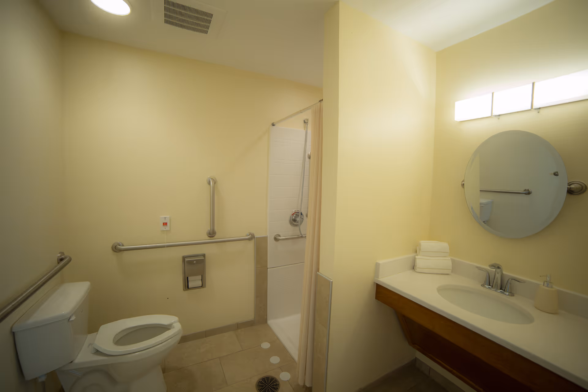 A clean, accessible bathroom with beige walls and tiled floor. The room features a toilet with grab bars on the walls, a walk-in shower with a curtain and grab bars, and a vanity with a sink, faucet, soap dispenser, a stack of folded towels, and a round mirror above it. A bright light fixture is mounted above the mirror.