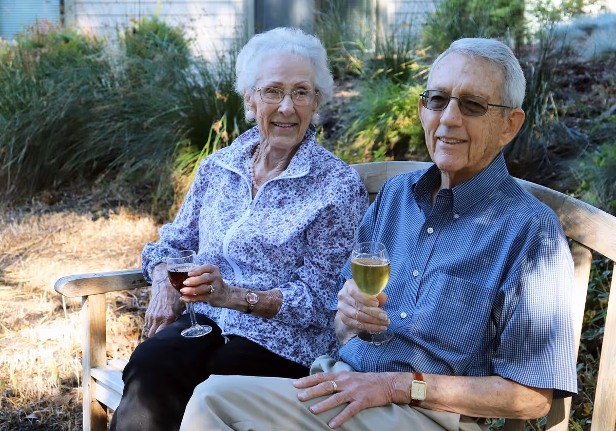An elderly woman and man sitting on a wooden bench outdoors, each holding a glass of wine. The woman is wearing glasses, a floral patterned jacket, and black pants, while the man is wearing glasses, a blue checkered shirt, and light-colored pants. They are surrounded by greenery and natural outdoor elements.