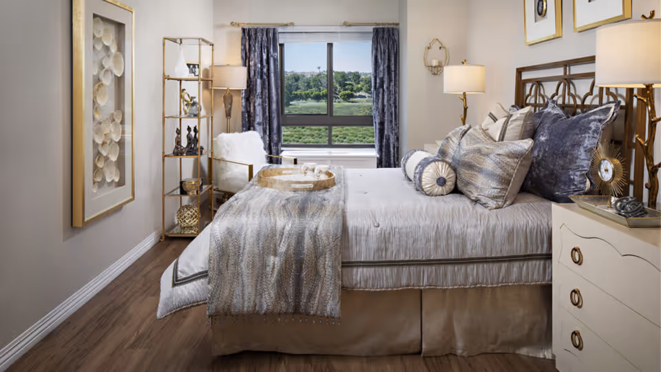 A well-decorated bedroom in a senior living facility featuring a large bed with multiple decorative pillows and a textured throw blanket. The room has a window with dark curtains overlooking greenery outside. There is a white armchair with a fluffy cushion, a gold metal shelving unit with decorative items, a nightstand with a lamp and clock, and framed artwork on the wall. The flooring is wood and the walls are painted a light neutral color.