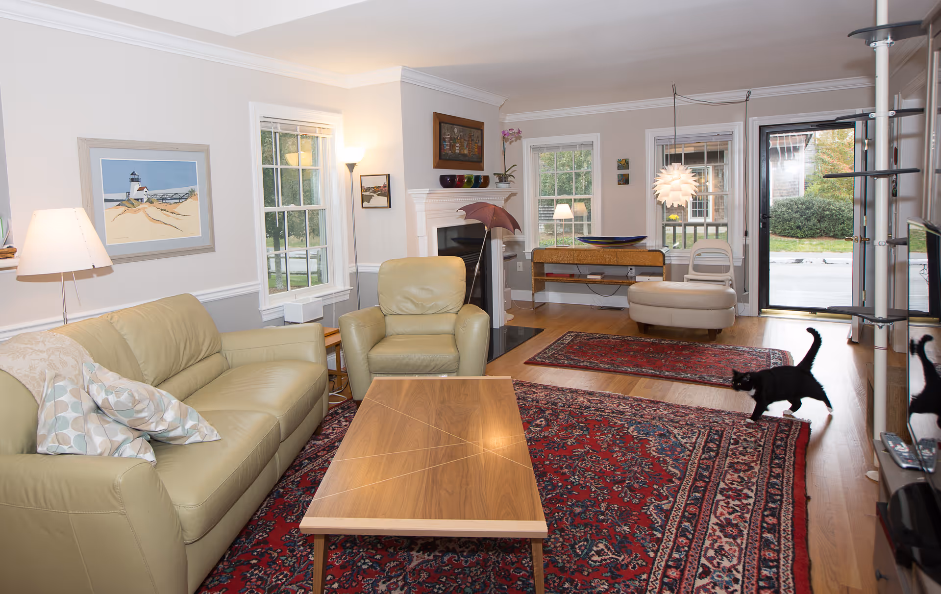A cozy living room with beige leather sofa and armchair, a wooden coffee table, and two red patterned rugs on a hardwood floor. The room has white walls with windows letting in natural light, a fireplace with decorative items on the mantel, a modern hanging light fixture, and a black cat walking near the entrance door.