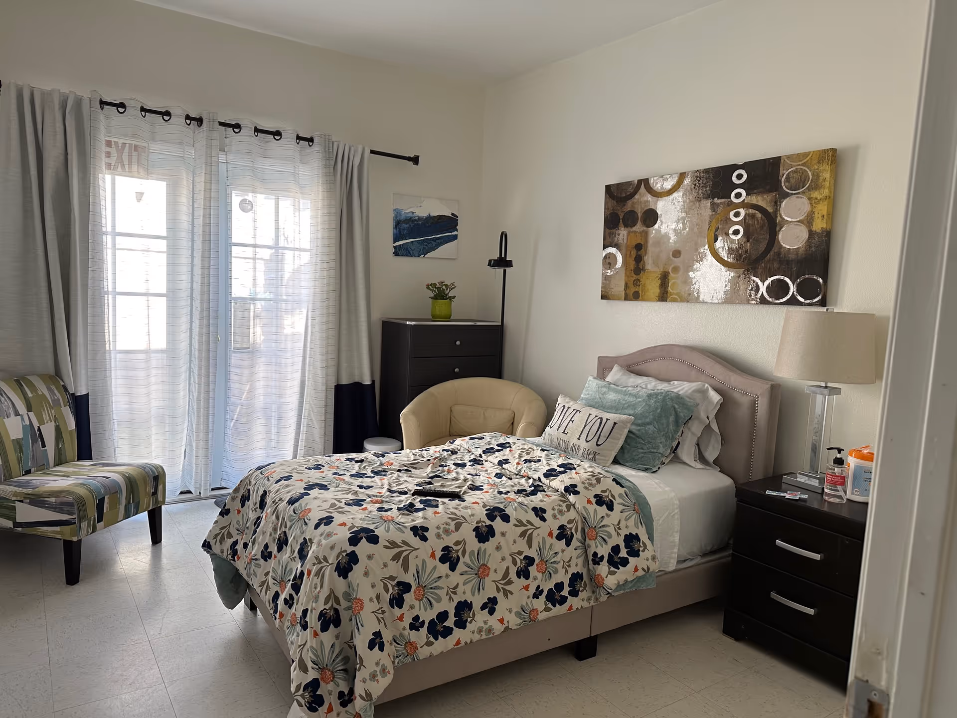 A cozy bedroom with a bed covered in a floral patterned comforter and multiple pillows, including one with the text 'LOVE YOU TO THE MOON AND BACK'. There is a beige armchair and a patterned chair near a window with sheer white curtains and gray drapes. A dark wooden dresser and nightstand with a lamp, hand sanitizer, and wipes are also visible. A modern abstract painting hangs above the bed.