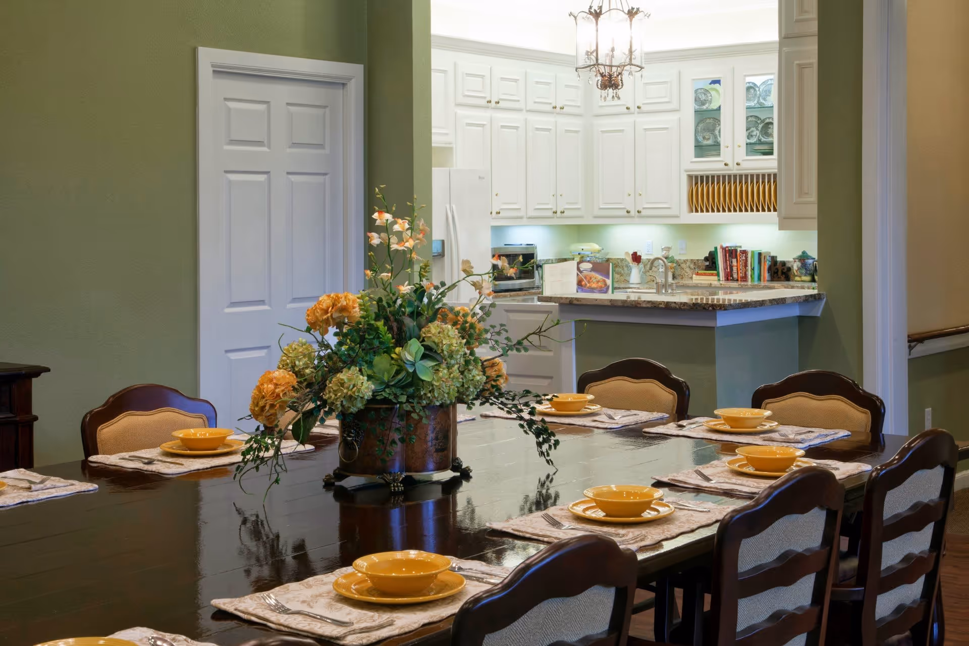 A dining room with a large dark wooden table set for eight people with yellow bowls and plates on beige placemats. A floral centerpiece with orange and green flowers is in the middle of the table. In the background, there is a kitchen with white cabinets, a countertop, and various kitchen items including a refrigerator and cookbooks.