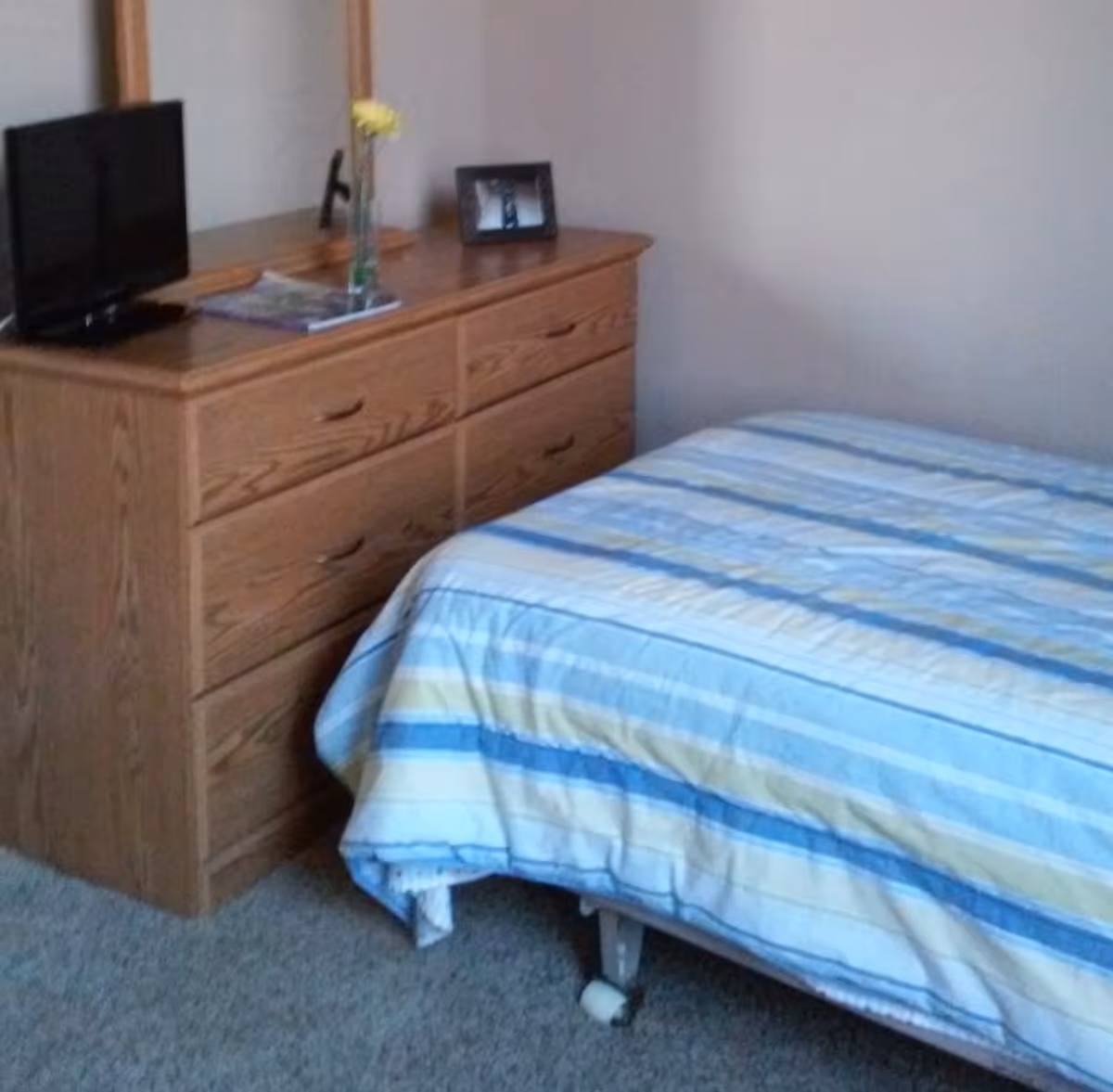 A bedroom with a bed covered in a striped blue, yellow, and white comforter. Next to the bed is a wooden dresser with six drawers, a small flat-screen TV, a framed photo, a magazine, and a vase with a yellow flower on top. The room has light-colored walls and carpeted flooring.