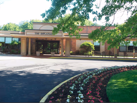 Front exterior view of The Esplanade at Palisades building with a covered entrance, surrounded by trees and a curved flower bed with red and white flowers.