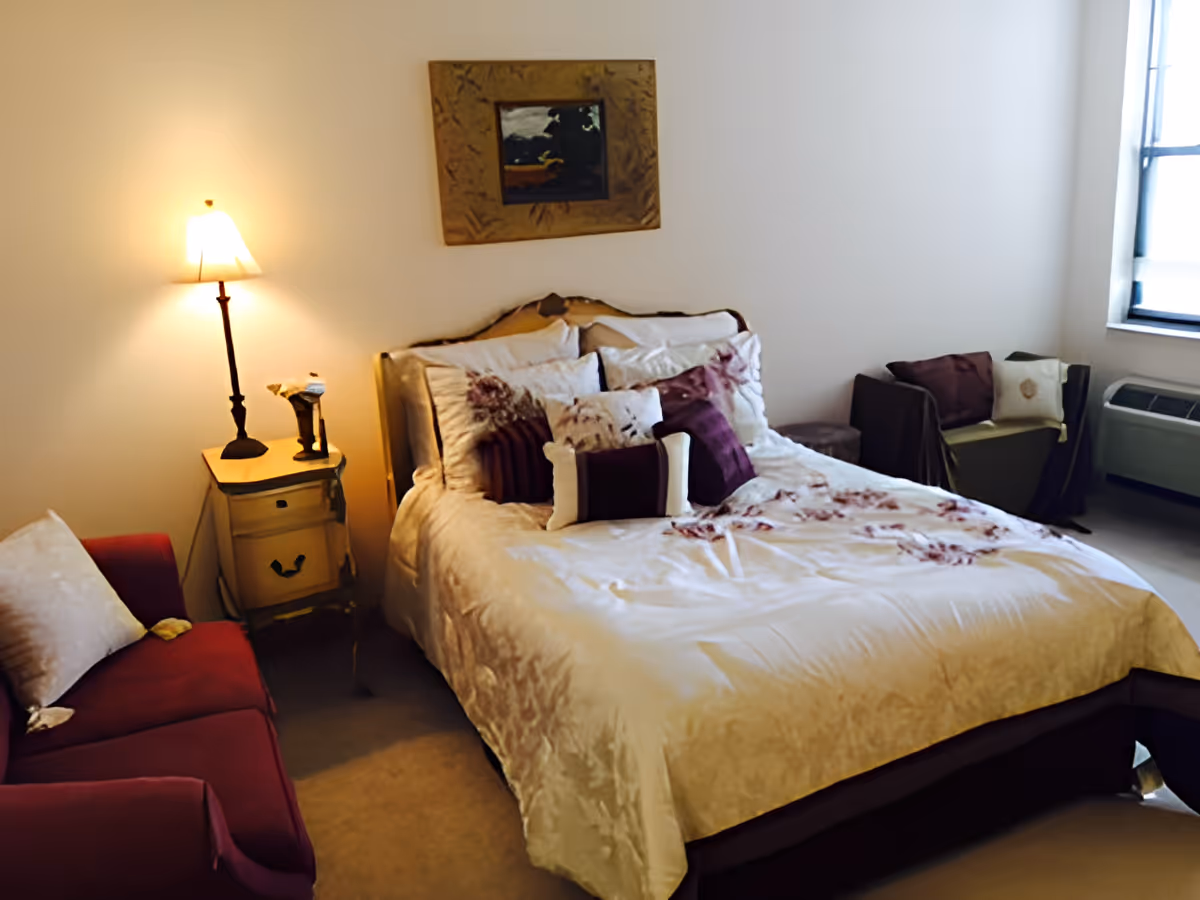 A cozy bedroom featuring a bed with cream and purple floral bedding and multiple pillows. To the left of the bed is a small wooden nightstand with a lamp and decorative items. A red armchair with a light-colored pillow is positioned near the nightstand. On the right side, there is a small seating area with a chair and pillows near a window with natural light coming through. A framed painting hangs on the wall above the bed.