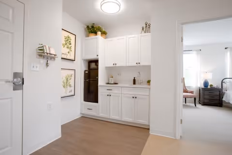 A bright interior space featuring a small kitchenette with white cabinets, a black refrigerator, and a countertop with a sink. There are decorative plants and framed botanical prints on the walls. To the right, an open doorway leads to a bedroom with a bed, nightstand, lamp, and a chair near a window with white curtains.