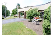 Exterior view of Caring Heights Community Care and Rehabilitation Center showing the building entrance with a covered drop-off area, a paved driveway, green lawn, trees, and a decorative white cart filled with flowers near the entrance.