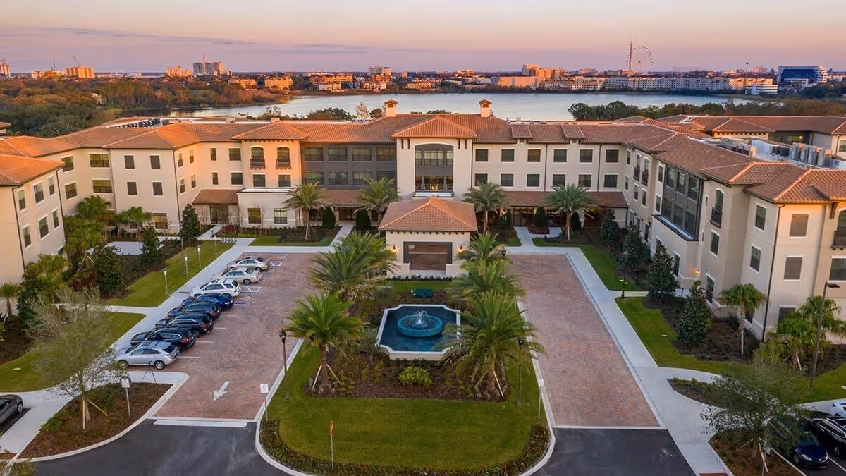 Aerial view of HarborChase of Dr. Phillips senior living facility showing a large, U-shaped building with a tiled roof surrounding a central courtyard with a fountain and palm trees. There is a parking area with cars on the left side, and a lake and cityscape are visible in the background during sunset.