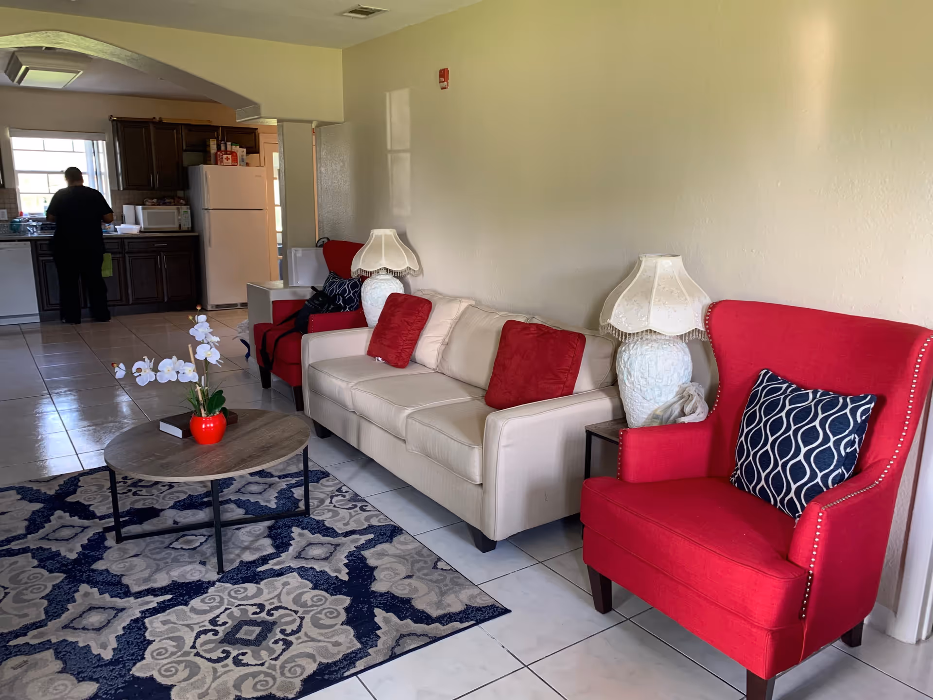 A living room area with a beige sofa adorned with red pillows, flanked by two red armchairs with patterned cushions. There are two white decorative lamps on side tables next to the sofa. A round wooden coffee table with a small red vase holding white flowers sits on a blue and gray patterned rug. In the background, a person is standing in a kitchen area with dark wood cabinets, a white refrigerator, and a microwave.
