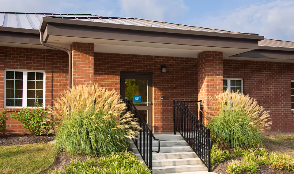 Entrance to a brick building with a small set of concrete stairs leading up to a glass door. The entrance is flanked by tall ornamental grasses and small shrubs, with black metal railings on both sides of the stairs. The building has white-framed windows and a metal roof under a partly cloudy sky.