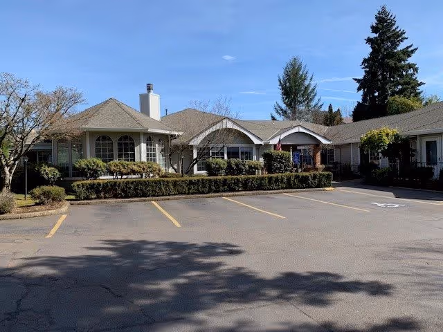 Exterior view of a single-story building with a gray roof and beige walls, surrounded by trimmed bushes and trees under a clear blue sky. There is an empty parking lot with marked spaces in front of the building.