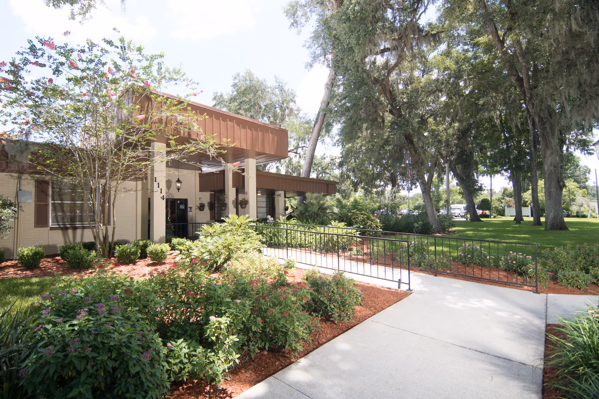 Exterior view of Brooksville Healthcare Center showing the entrance with a covered walkway, surrounded by landscaped greenery, bushes, and trees. A concrete path with a black metal railing leads to the entrance under a brown awning.