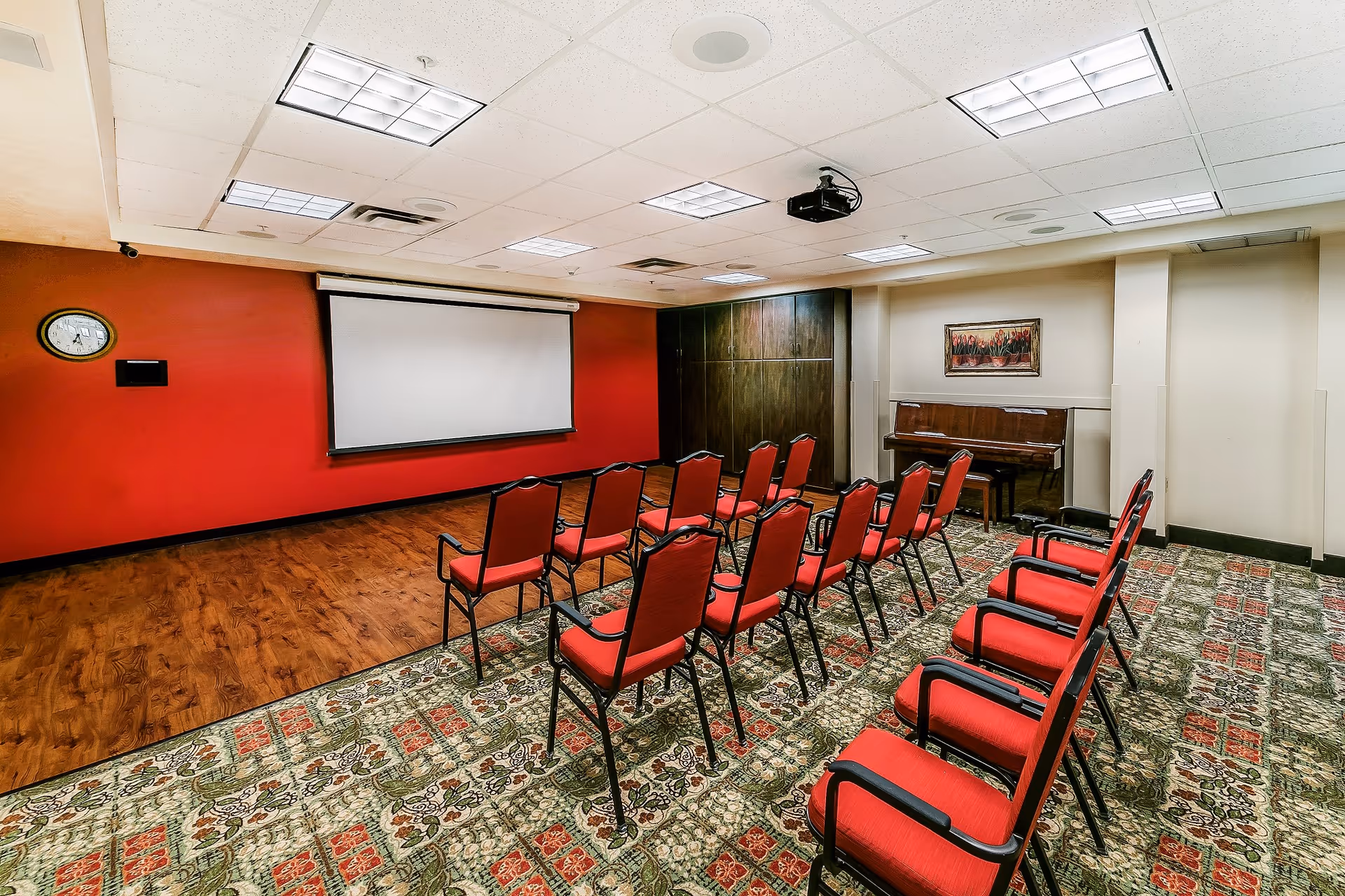 A small conference or presentation room with two rows of red cushioned chairs facing a white projection screen mounted on a red wall. The room has a patterned carpet, a wooden floor section near the screen, a piano against the far wall, and a framed painting above the piano. The ceiling has multiple fluorescent lights and a projector mounted in the center.