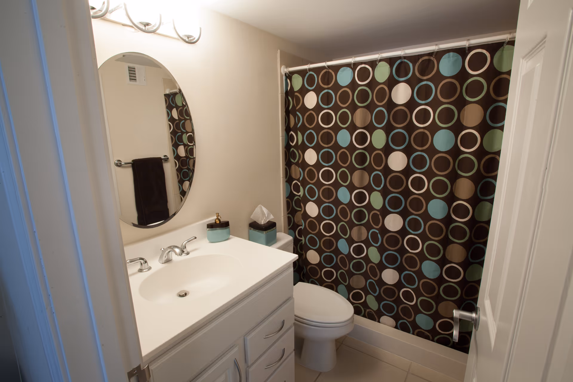 A small bathroom with a white vanity and sink, an oval mirror above the sink, a toilet next to the vanity, and a shower with a curtain featuring a pattern of blue, green, brown, and white circles.