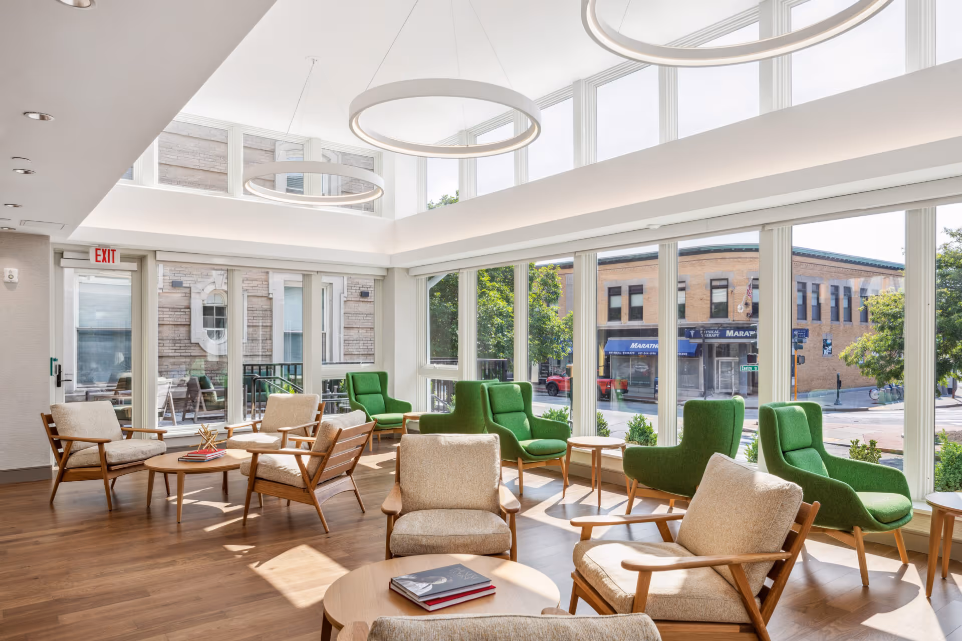 A bright and modern common area with large floor-to-ceiling windows letting in natural light. The room features a mix of beige cushioned armchairs and green upholstered chairs arranged around small wooden tables on a wooden floor. Circular pendant lights hang from the ceiling, and outside the windows, a street with buildings and trees is visible.