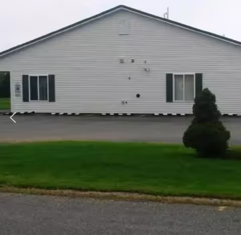 Exterior view of a single-story white building with green shutters on two windows, a paved driveway in front, and a small bush on a grassy area to the right.
