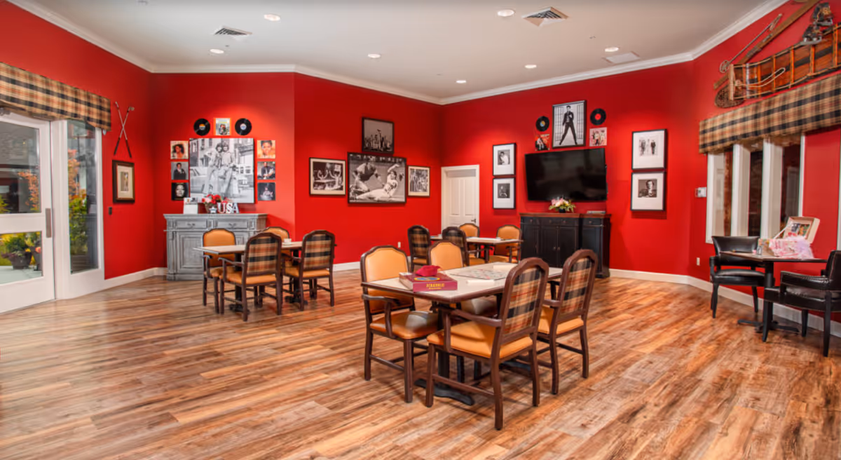 A bright room with red walls and wooden flooring featuring several tables and chairs arranged for group activities. The walls are decorated with framed black and white photos, vinyl records, and a mounted sled. There is a flat-screen TV mounted on one wall and large windows with plaid valances allowing natural light to enter. The room appears to be a communal space for socializing or activities.