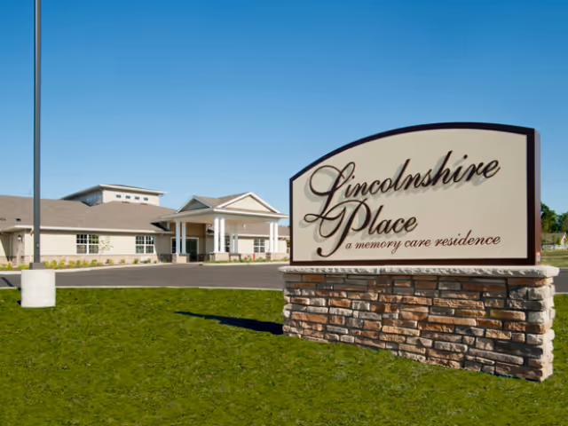 Exterior view of Lincolnshire Place memory care residence building with a large sign in the foreground displaying the facility name. The building has a light-colored facade, a covered entrance with columns, and a clear blue sky above.