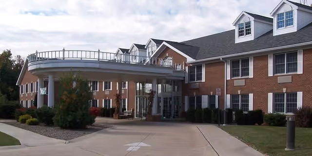 Exterior view of a brick assisted living facility building with multiple windows and dormers on the roof. There is a covered entrance supported by white columns, a driveway leading up to the entrance, and landscaping with bushes and trees around the building.