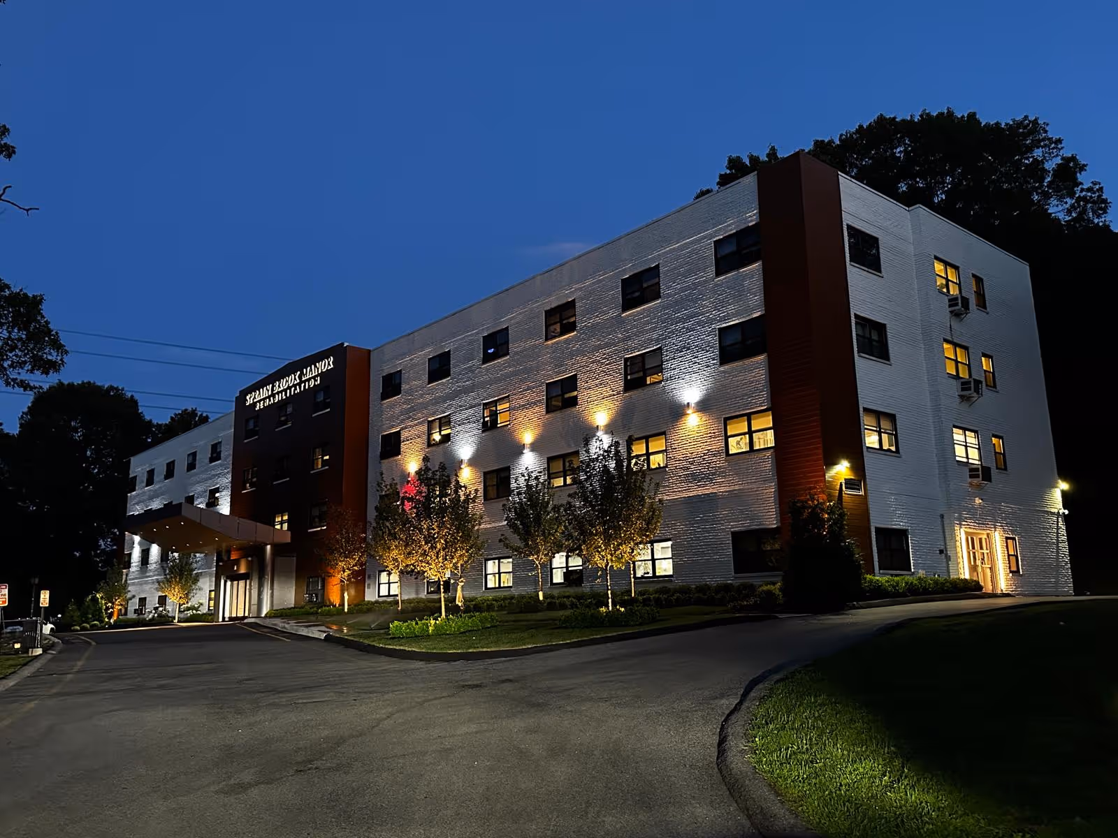 Exterior view of Sprain Brook Manor Rehab building at dusk, showing a three-story white and brown brick structure with illuminated windows and outdoor lights, surrounded by trees and a driveway.