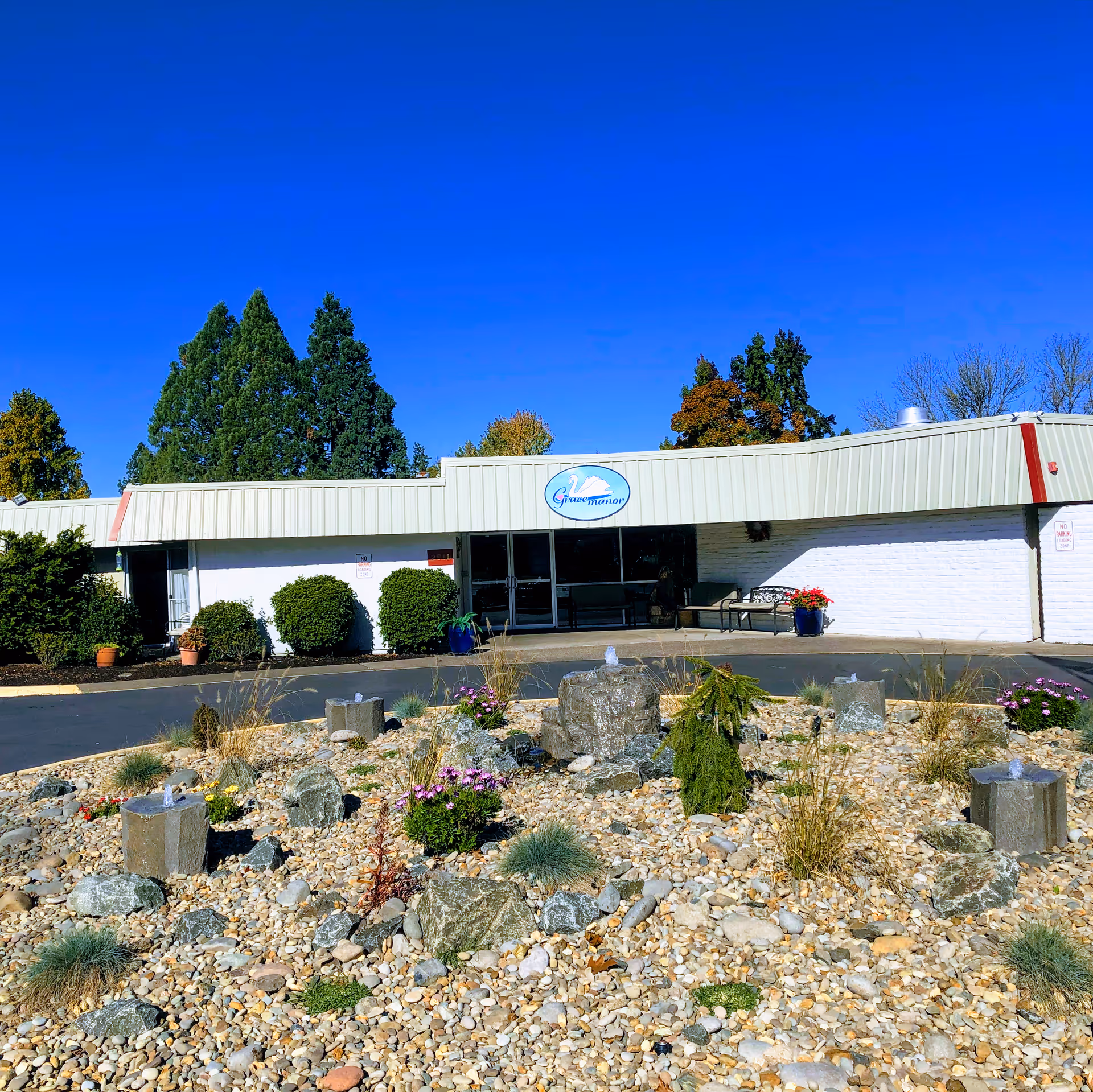 Exterior view of Grace Manor Senior Living facility with a white building, a sign displaying the facility's name, a rock garden with small plants and flowers in the foreground, and tall green trees in the background under a clear blue sky.