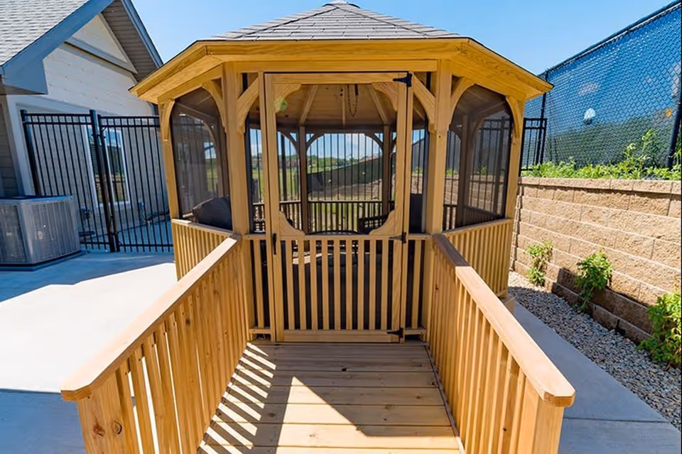 A wooden gazebo with screened windows and a small gate at the entrance, located outdoors next to a building and a fenced area, with a clear blue sky above.