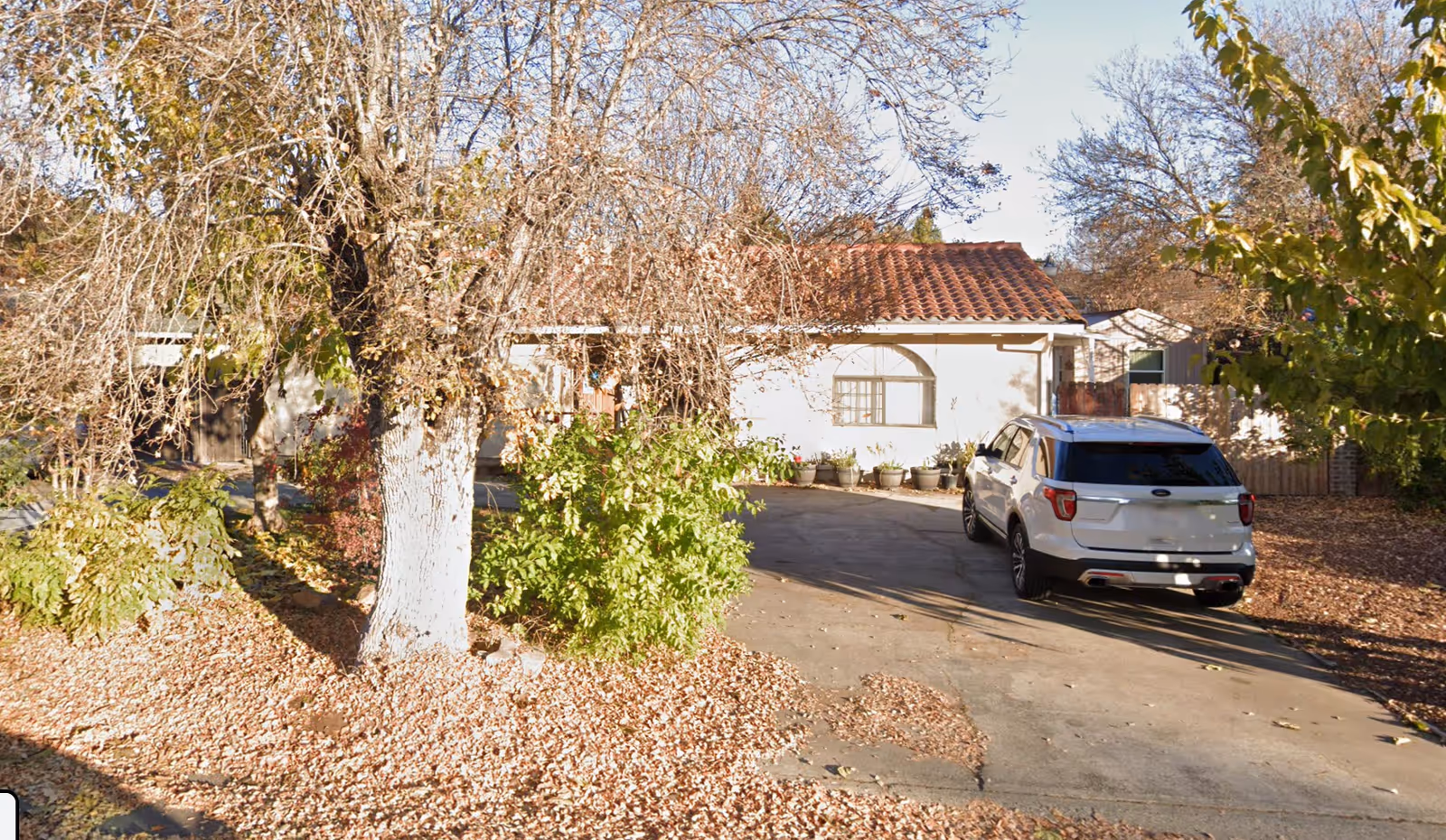 A single-story house with a tiled roof partially obscured by leafless trees and bushes. A white SUV is parked on the driveway in front of the house. The ground is covered with fallen leaves, indicating an autumn setting.