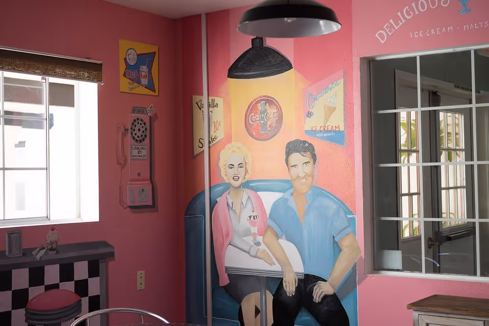 Interior corner of a room with pink walls featuring a mural of a man and woman sitting in a retro diner booth. The mural includes vintage-style advertisements for Coca-Cola, ice cream, and milkshakes. A window with white framing and a bamboo shade is on the left wall, and a small table with a checkered pattern and a red stool is partially visible. A black hanging lamp is above the mural.
