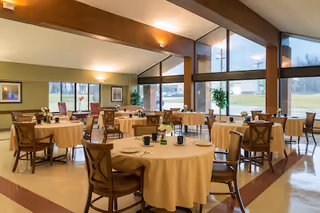 A spacious dining room with round tables covered in beige tablecloths, each set with cups and small flower arrangements. Wooden chairs surround the tables. Large windows let in natural light and offer a view of the outdoors. The ceiling has wooden beams and wall-mounted lights provide additional illumination.
