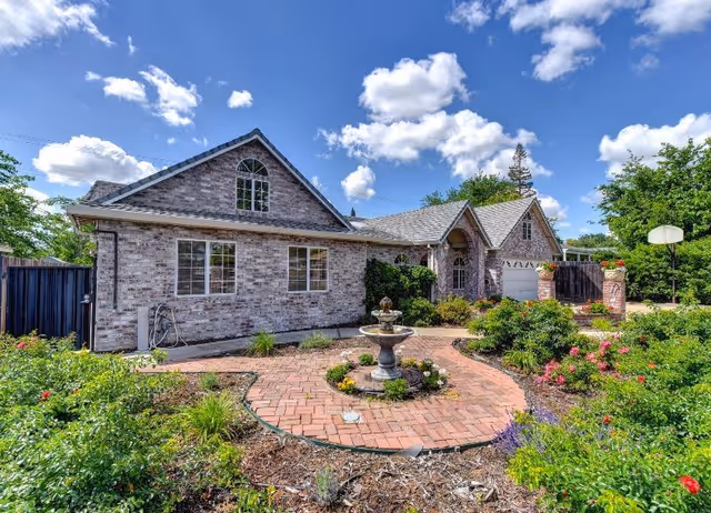 Exterior view of a single-story brick building with multiple windows and a gabled roof under a partly cloudy blue sky. In front of the building is a circular brick pathway surrounding a small fountain, with various green shrubs and flowering plants in the garden area.