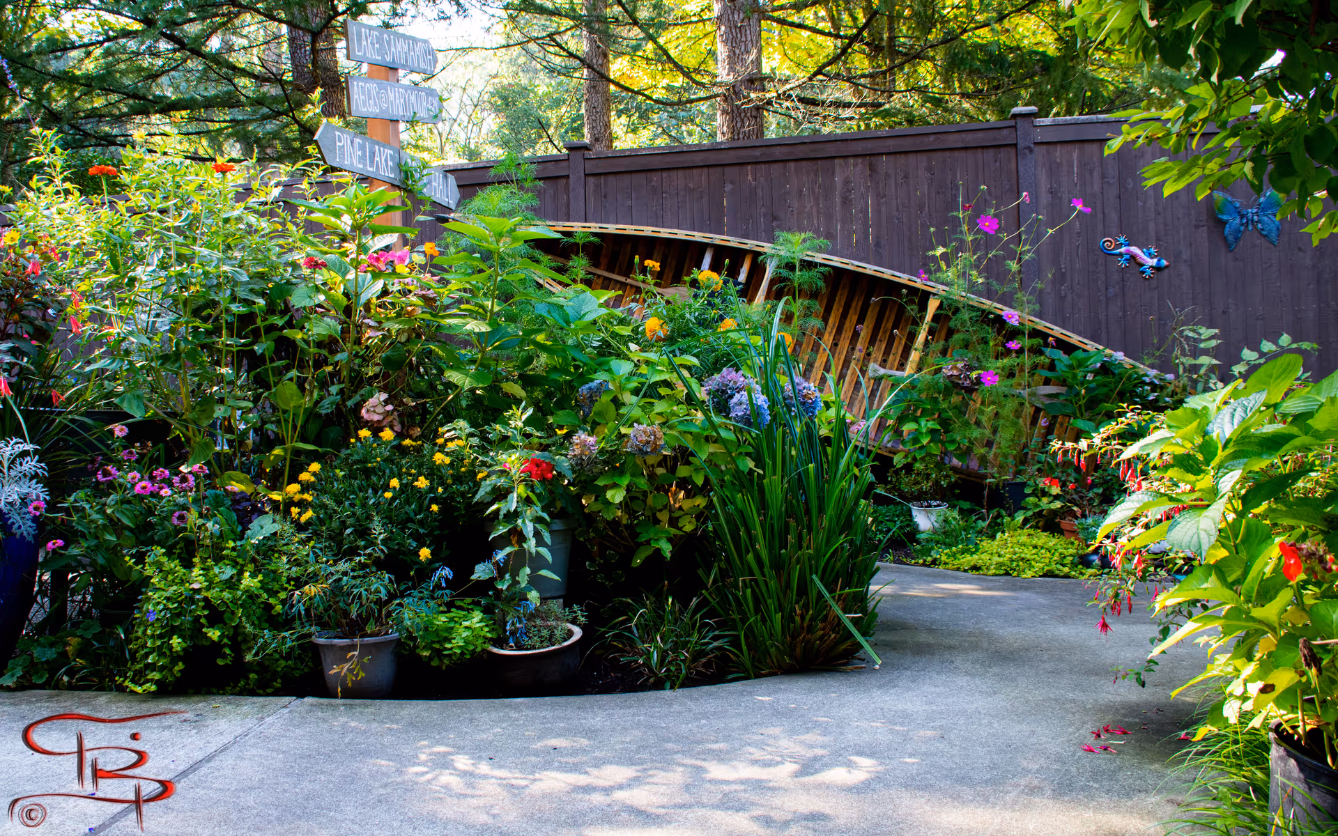 A vibrant garden area with various colorful flowers and green plants surrounding a concrete pathway. A wooden fence is in the background with a wooden boat leaned against it. There is a wooden signpost with multiple directional signs pointing to different locations such as Lake Sammamish, Aegis Marymoor, Pine Lake, and Hall.