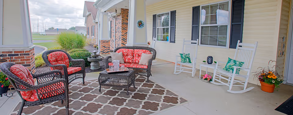Outdoor covered patio area with wicker furniture including a loveseat and two chairs with red floral cushions, a coffee table, two white rocking chairs with green leaf patterned pillows, a small white side table with a decorative item, and potted plants on the concrete floor. The patio is adjacent to a yellow building with windows and black shutters.