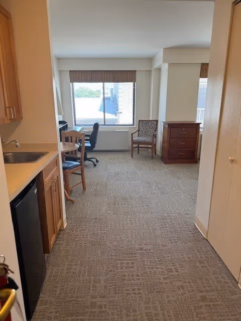Interior view of a senior living facility room with a kitchenette on the left, including a sink, mini refrigerator, and cabinets. The room has carpeted flooring and a window at the far end with a chair and a small desk with a rolling office chair in front of it. There is also a wooden dresser and another chair near the window.