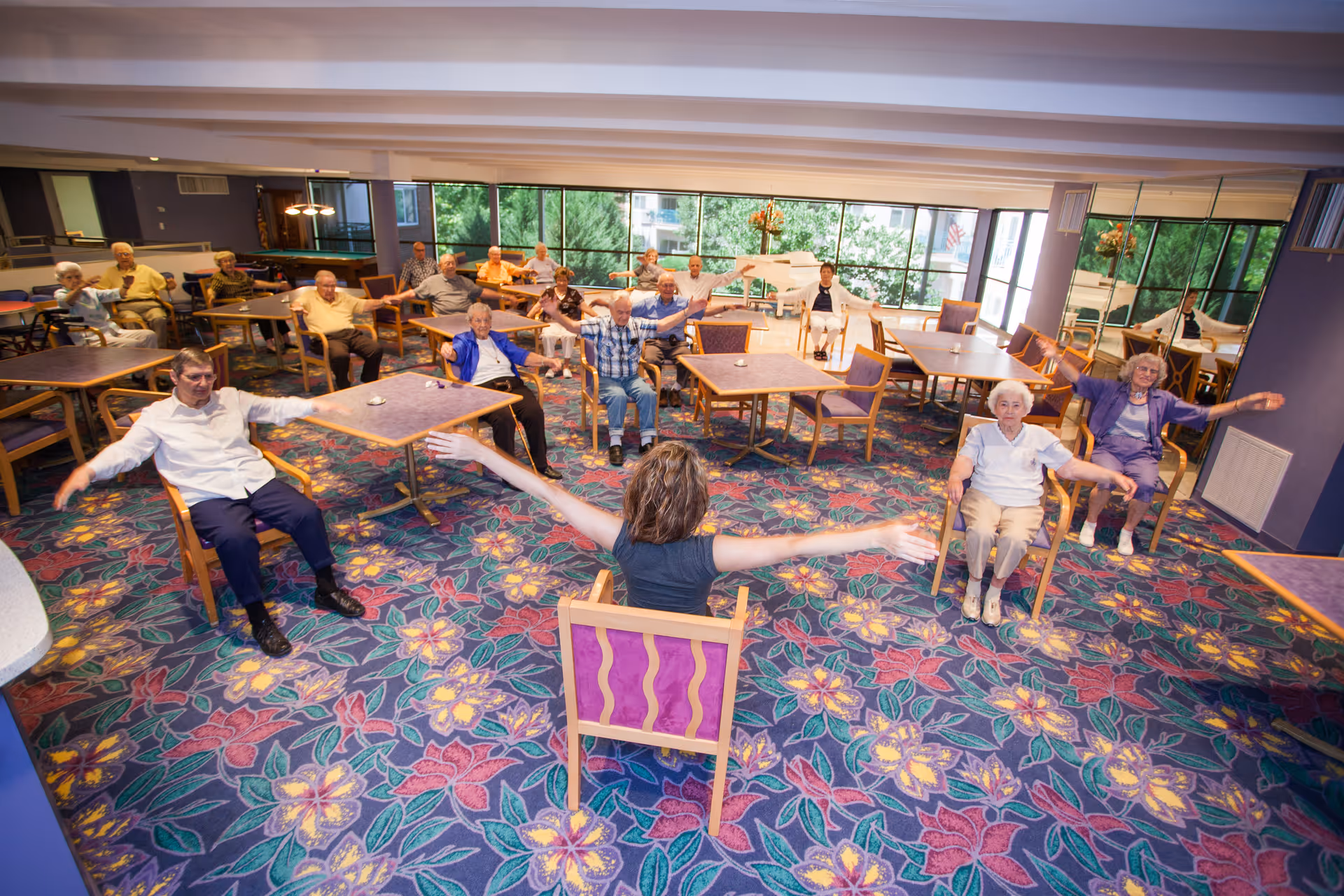 A group of elderly people seated in chairs arranged in a spacious room with large windows, participating in a seated exercise session led by an instructor facing them with arms extended.