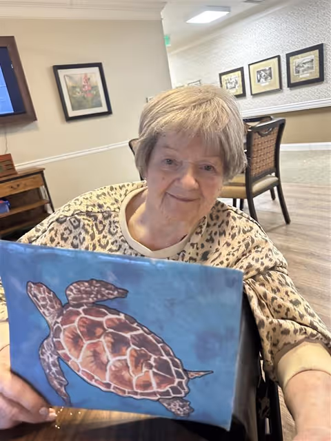 An elderly woman with short gray hair wearing a leopard print top is sitting at a table in a well-lit room. She is holding up a blue canvas with a painted sea turtle. The room has framed pictures on the walls and wooden furniture.