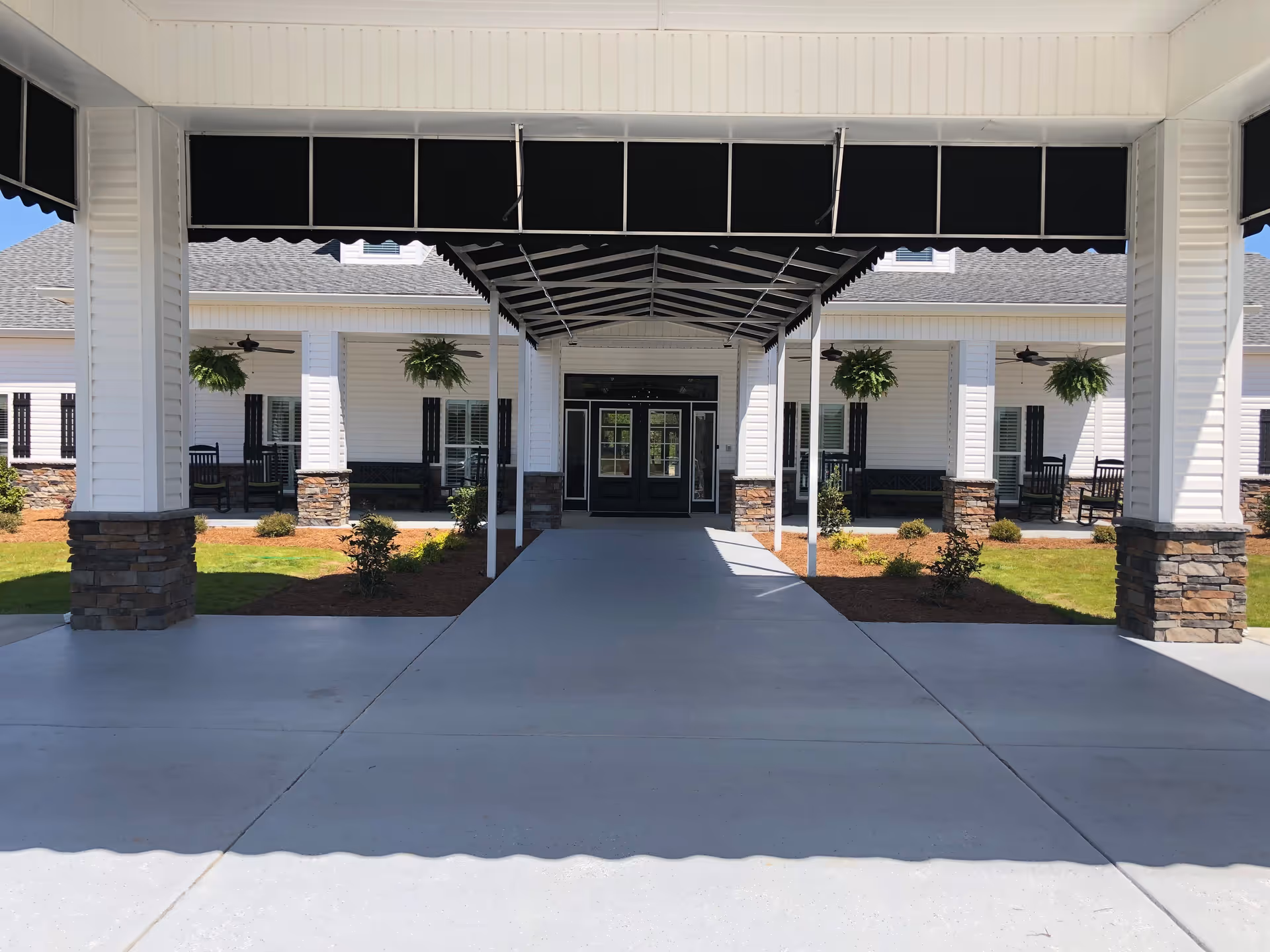 Covered driveway entrance leading to the front doors of a senior living facility with white siding, stone pillars, hanging plants, and rocking chairs on the porch.