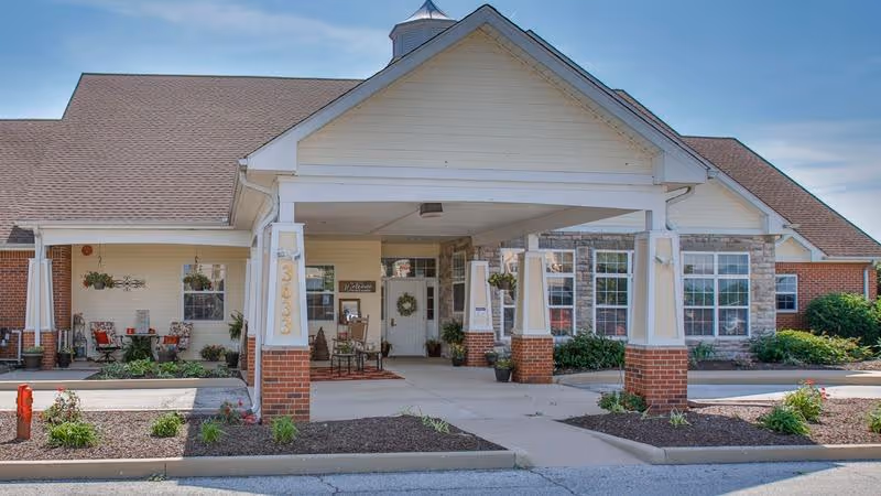 Front exterior view of a senior living facility with a covered entrance supported by white columns with brick bases. The building has a combination of beige siding and stone accents, with large windows and a welcoming seating area near the entrance. The sky is clear and blue.