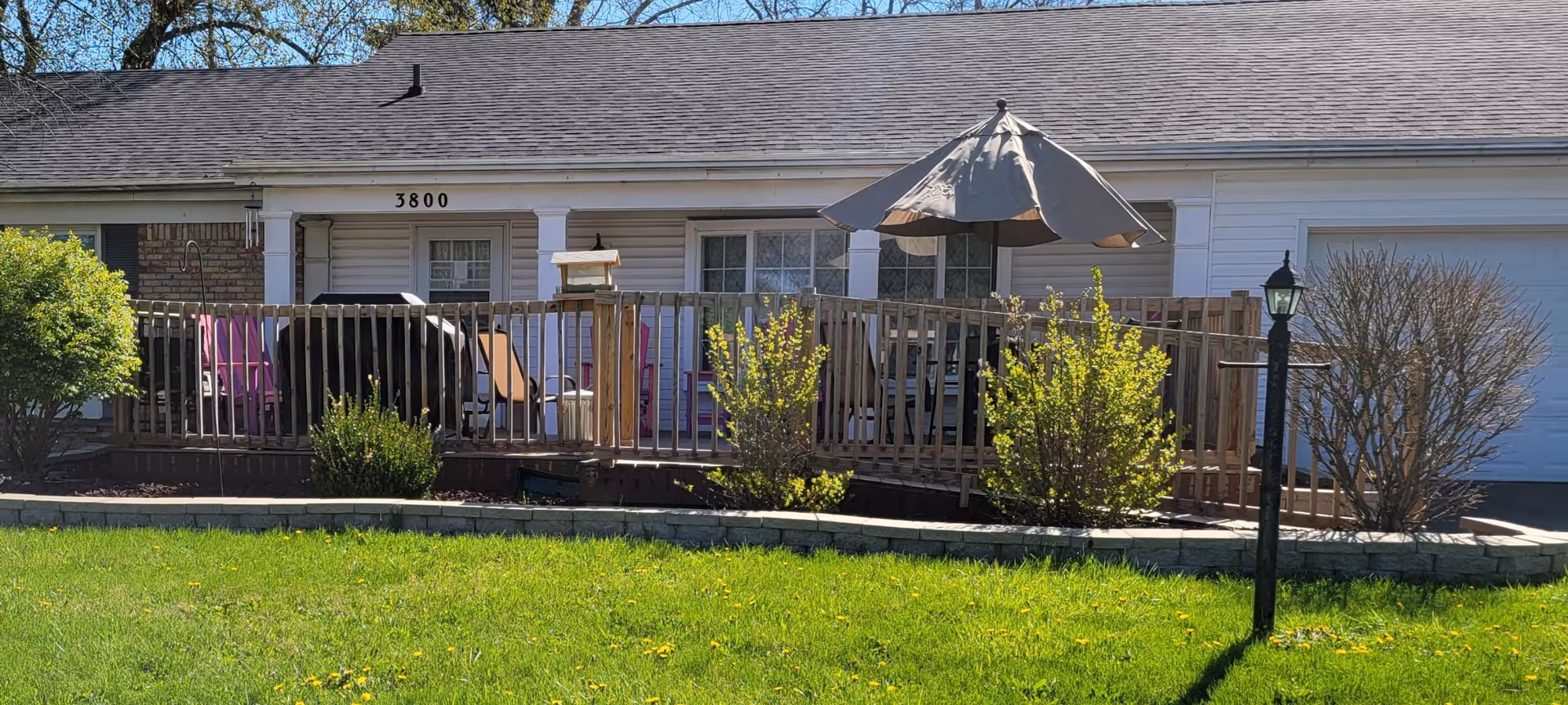 Front exterior view of a single-story residential building with white siding and a gray roof. The house number 3800 is visible above the front door. There is a wooden deck with a railing, outdoor furniture including chairs and a table with a large umbrella. Shrubs and a lamp post are in the landscaped area in front of the deck, and a green lawn is in the foreground under a clear blue sky.