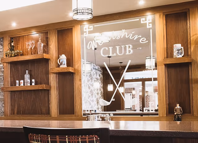 Interior view of a wooden bar area with shelves holding glassware and decorative items. A large mirror behind the bar displays the text 'Wealshire CLUB' with crossed golf clubs graphic. Pendant lights hang from the ceiling and a chair with a patterned back is partially visible in the foreground.