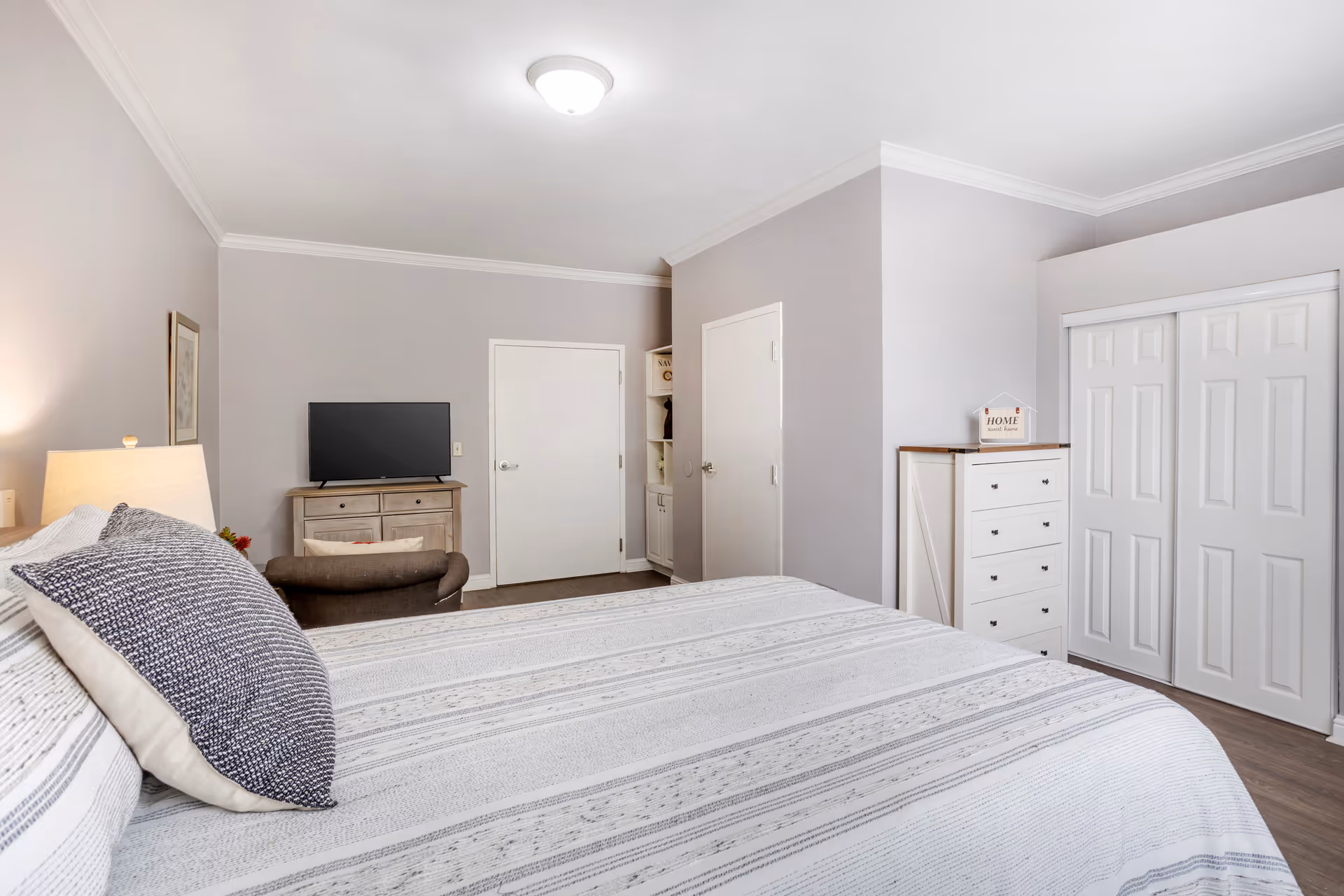 A bedroom with a large bed covered in a white and gray striped bedspread and pillows. There is a wooden dresser with a flat-screen TV on top, a small armchair, two white doors, a white chest of drawers with a decorative 'Home Sweet Home' sign, and a closet with sliding doors. The walls are painted light gray and the floor is wood.